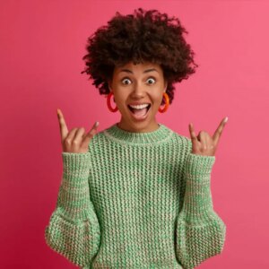happy carefree dark skinned rebellious young woman enjoys awesome music makes rock n roll gesture has fun on music festival or cool event wears casual jumper poses against pink wall | MXO Media Agency